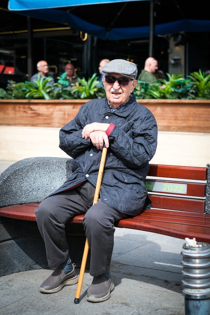 Senior man with cane sitting on a bench outdoors, wearing a cap and sunglasses.