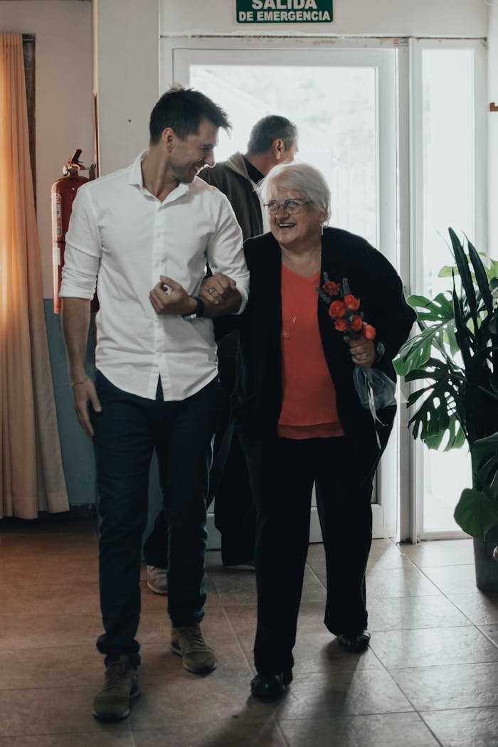 Senior woman arm in arm with a man, smiling and walking indoors with a bouquet of flowers.